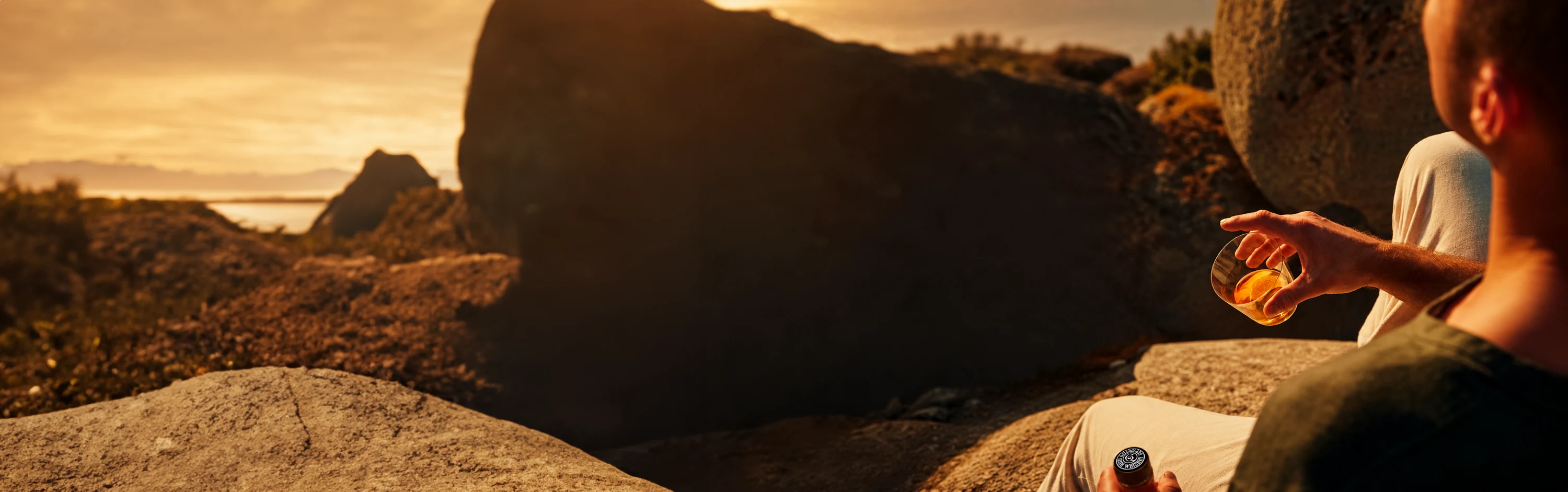 A man sits among sunlit coastal rocks at golden hour, holding a glass of whiskey and a bottle of Galloglass